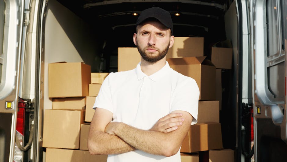 A man with a serious expression, wearing a white polo shirt and a black cap, stands inside an open moving van surrounded by numerous cardboard boxes of various sizes, some packed with clear plastic wr