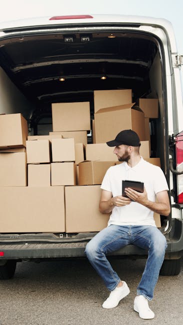 A man with a serious expression, wearing a white polo shirt and a black cap, stands inside an open moving van surrounded by numerous cardboard boxes of various sizes, some packed with clear plastic wr